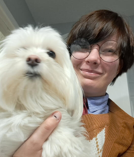 Clark, a younger person with straight, short, dark hair and wearing big, round glasses, a blue dress shirt, and a brown sweater and jacket holds his cream-colored, fluffy dog, Louie.